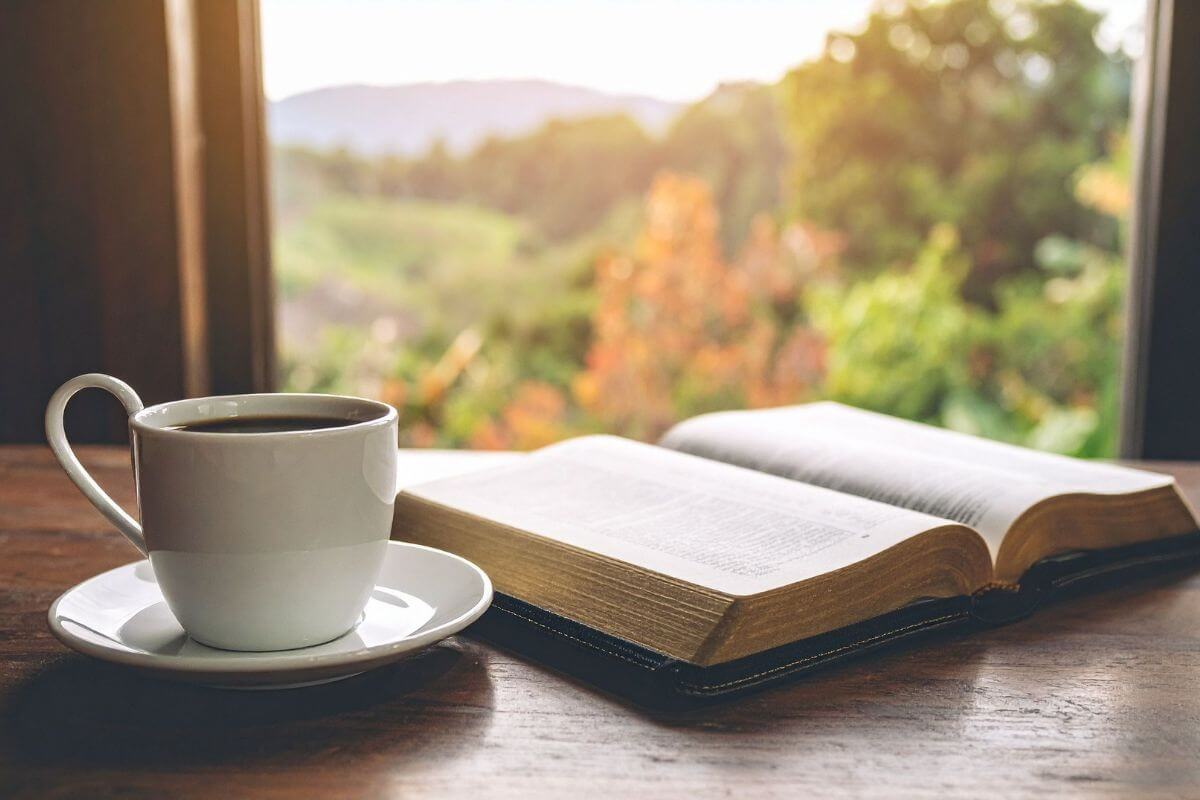 Open bible with a coffee mug beside it on a table by an open window representing spending time in God's word.