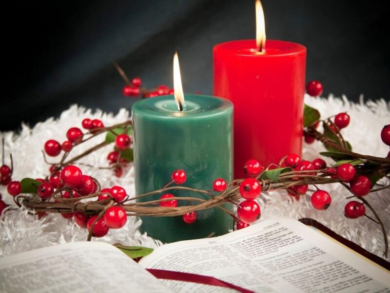 Two lit candles—one red and one green—surrounded by a wreath of red berries and green leaves, resting on a white textured surface beside an open Bible showing Luke 2:10. A peaceful Christmas scene symbolizing faith and reflection, perfect for posts about Christmas scriptures for hope and peace.