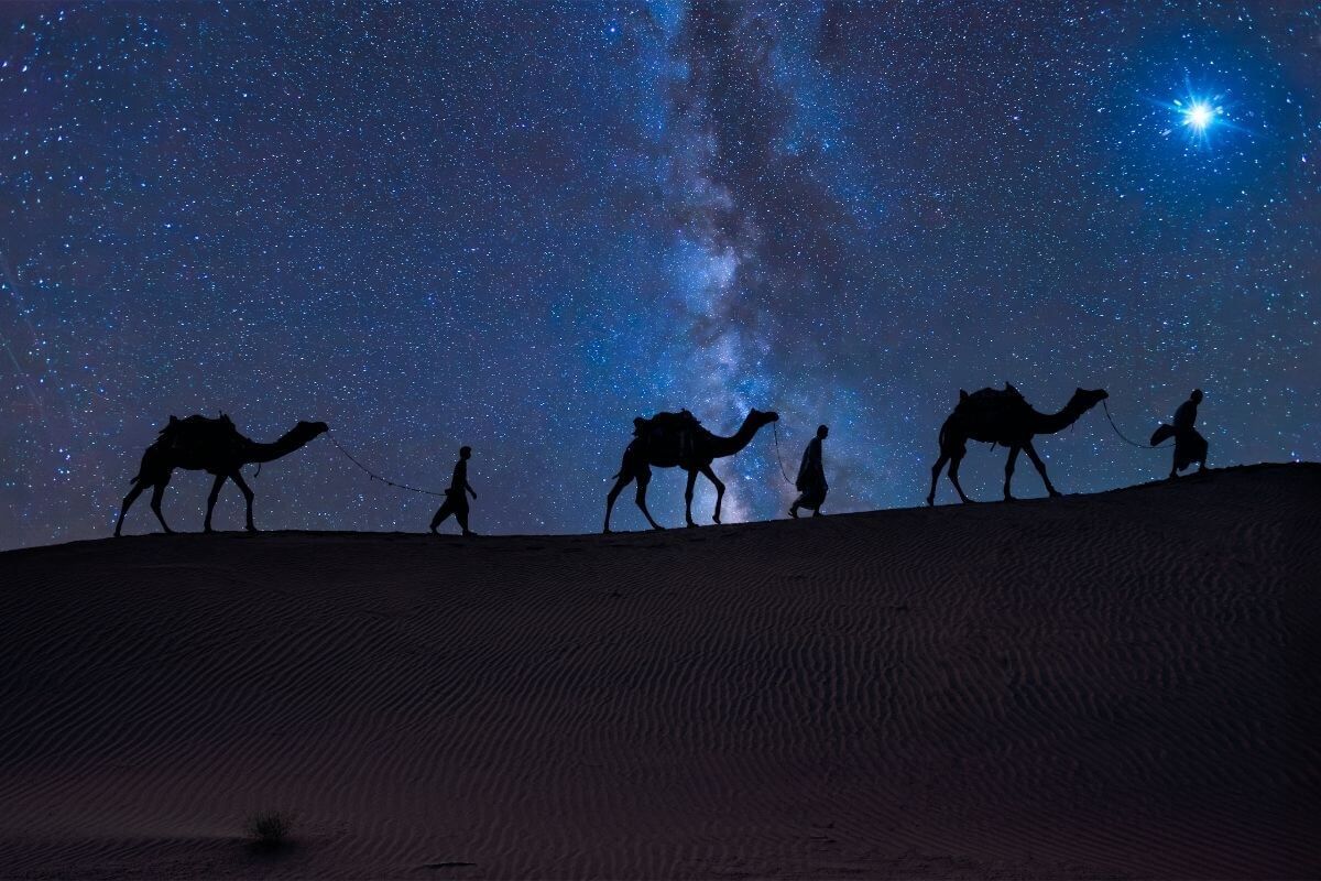 Silhouette of three people leading camels across a desert under a star‑filled night sky. The Milky Way glows brightly overhead, and a single radiant star shines near the horizon, symbolizing divine timing and the journey of faith as God sends His Son at the appointed time.