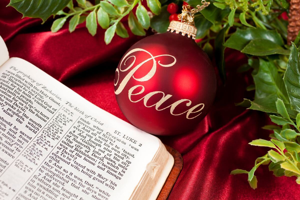 A bible opened to Luke chapter 2 on a decorated table with red Christmas ball with peace written on it and a green flower beside it.