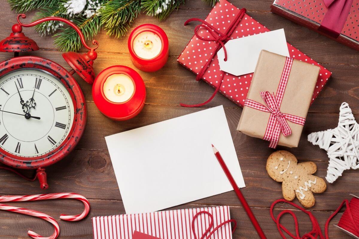 Table containing wrapped Christmas gifts; two red lit candles, red clock, a Christmas card and red pen laying over it.