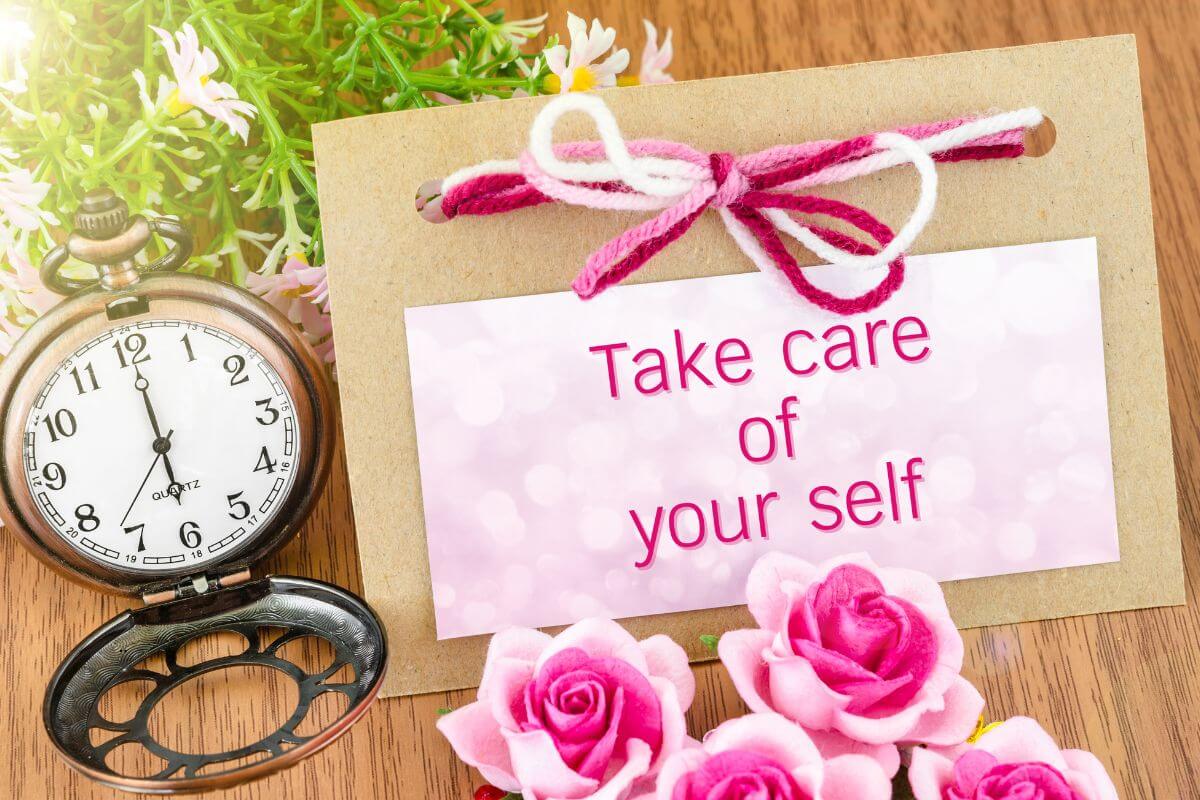 A vintage pocket watch sits open on a wooden surface beside a card tied with pink and white yarn that reads “Take care of yourself.” Pink roses and small flowers surround the card, creating a gentle reminder of self‑care when Christmas feels overwhelming.