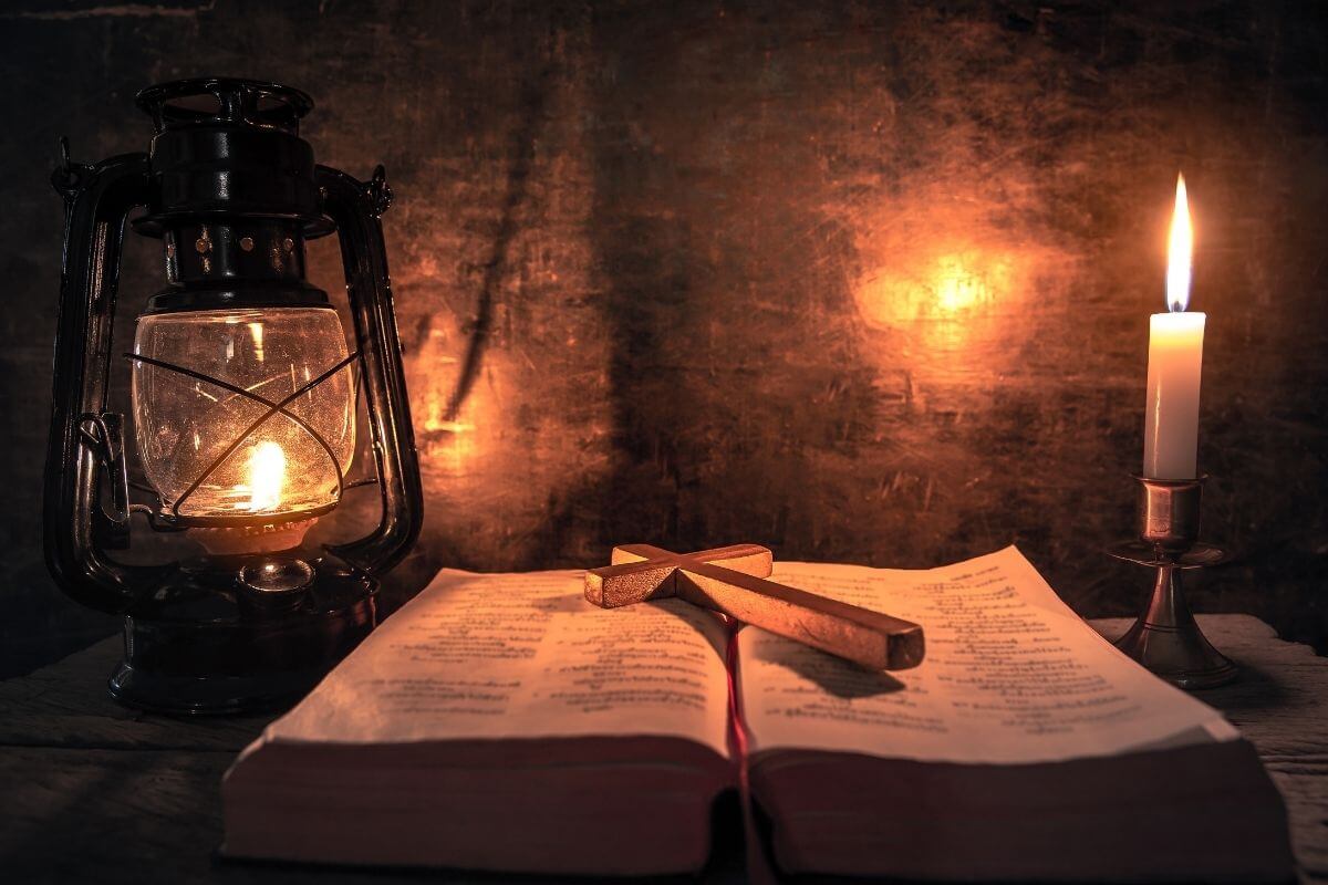 An open Bible rests on a wooden table with a wooden cross placed across its pages. A lantern and a candle glow softly on either side, casting warm light that symbolizes faith and remembrance of grace through Jesus. The peaceful scene illustrates Christmas scriptures for hope and peace, inviting reflection and gratitude.