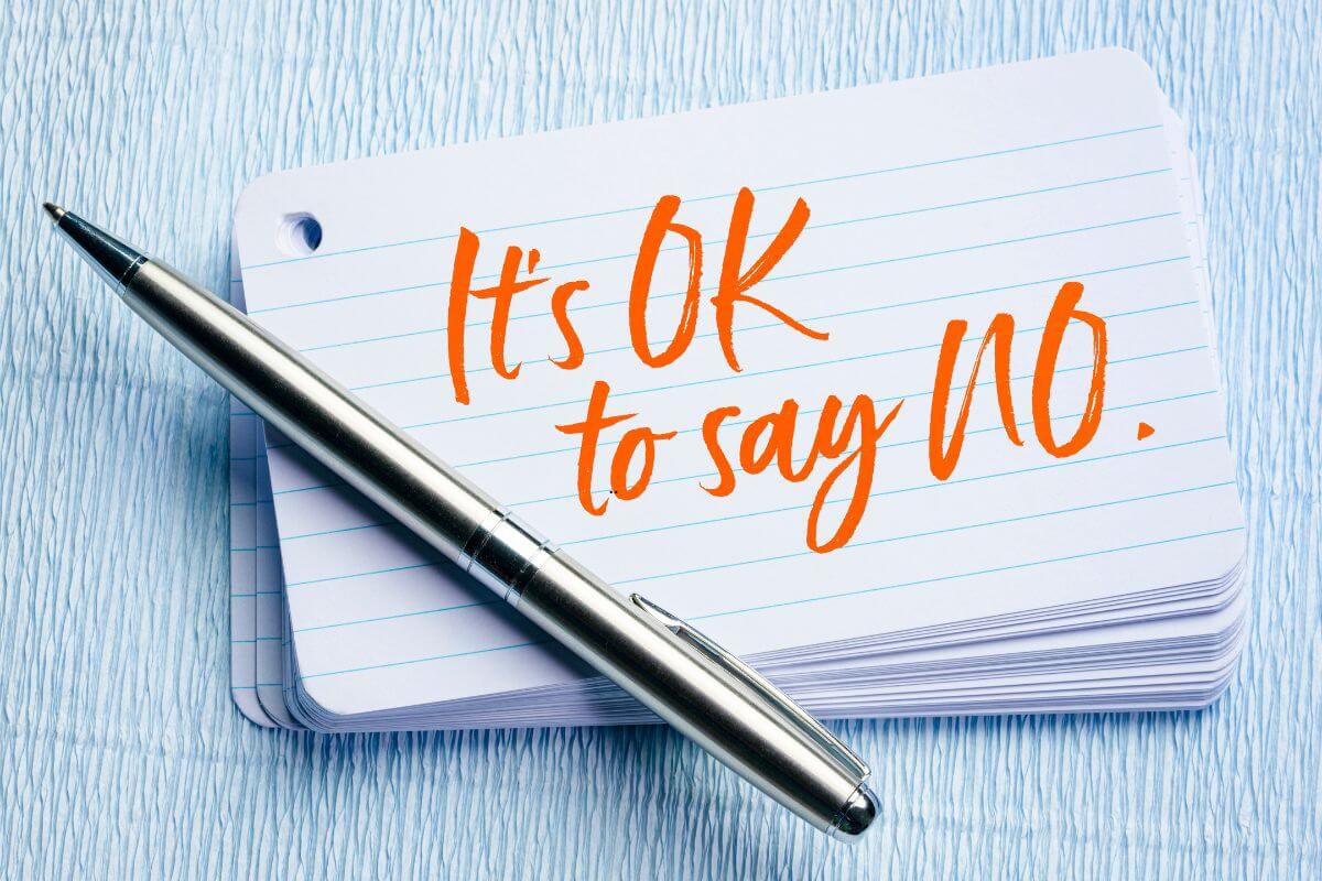 A silver pen rests diagonally across a stack of white lined index cards on a light blue textured surface. The top card has bold orange handwritten text that reads “It’s OK to say NO,” symbolizing healthy boundaries and self‑care