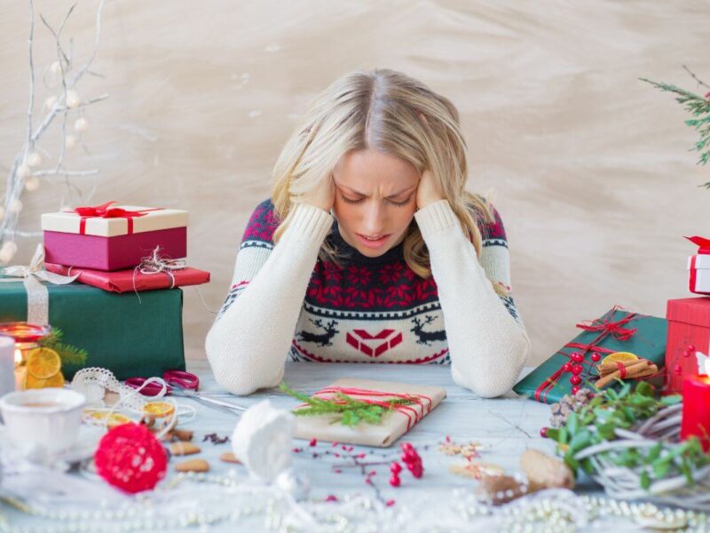 Woman sitting at a table covered with Christmas decorations and wrapped gifts, wearing a festive sweater with reindeer and snowflake patterns. She rests her head in her hands, looking stressed amid holiday preparations, capturing the emotion and tension that arise when Christmas feels overwhelming.