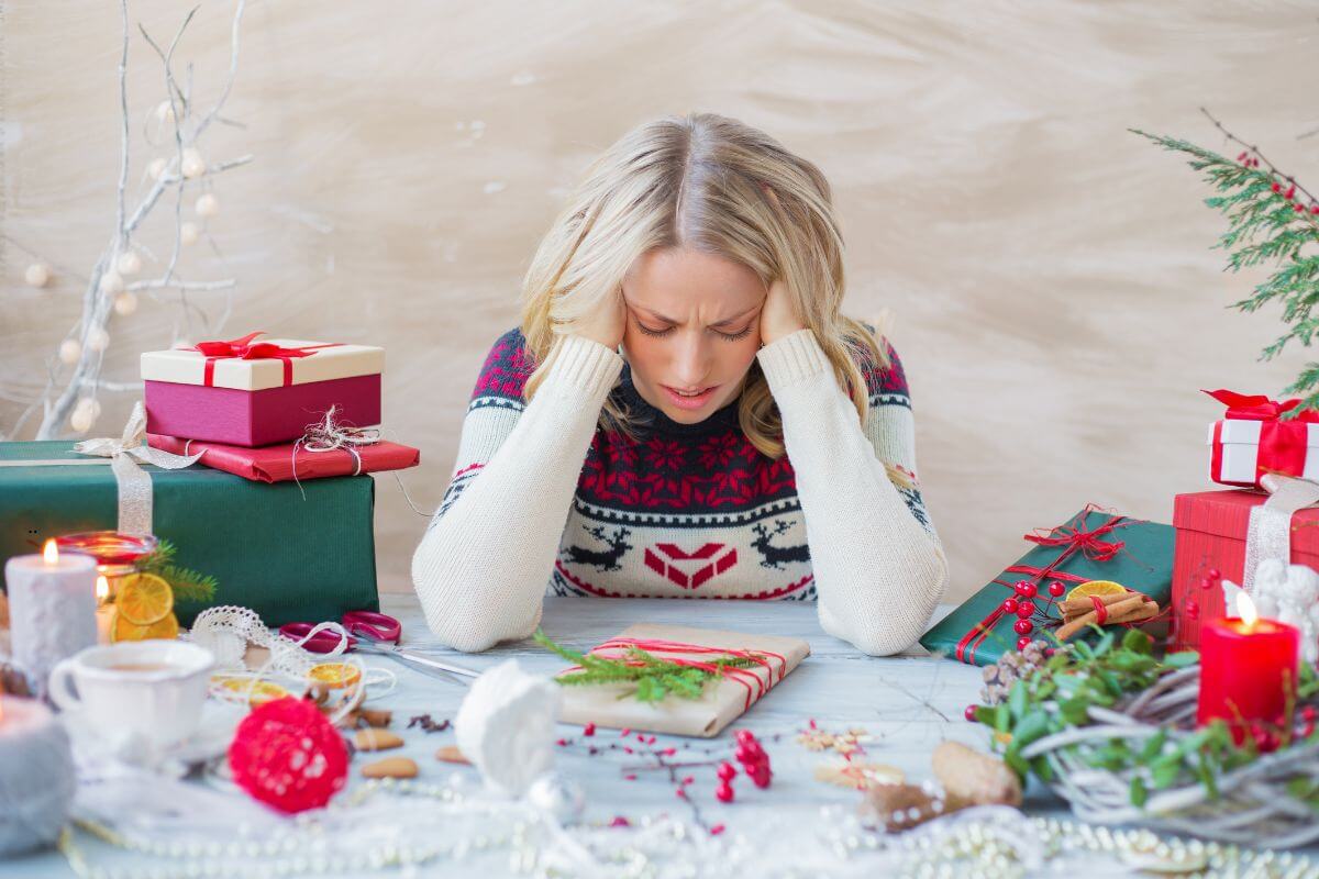 Woman sitting at a table covered with Christmas decorations and wrapped gifts, wearing a festive sweater with reindeer and snowflake patterns. She rests her head in her hands, looking stressed amid holiday preparations, capturing the emotion and tension that arise when Christmas feels overwhelming.