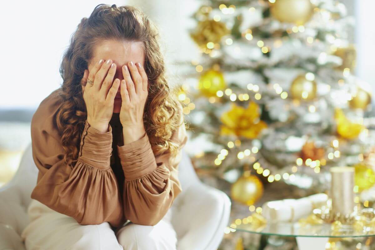 Woman sitting indoors with her face buried in her hands, wearing a light brown blouse and white pants. A decorated Christmas tree with gold ornaments and warm lights glows softly in the background, capturing the emotional weight and stress when Christmas feels overwhelming.