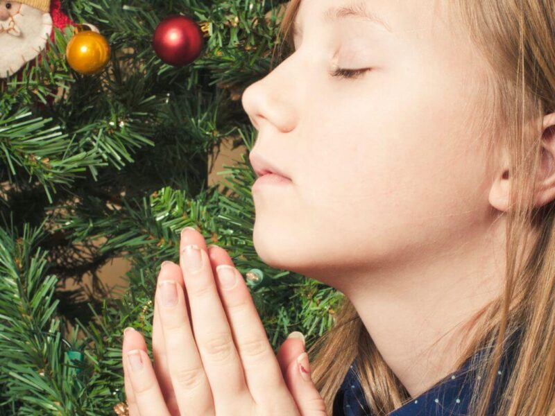 Young white lady with eyes closed and hands in prayer in front of a Christmas tree.