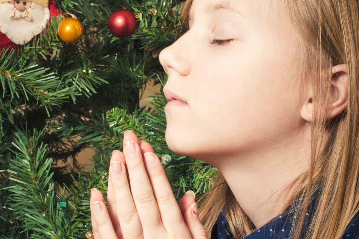 Young white lady with eyes closed and hands in prayer in front of a Christmas tree.